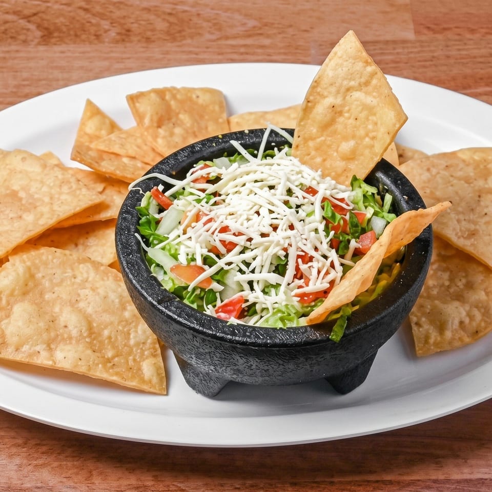 Black bowl of guacamole topped with shredded cheese, diced tomatoes, and onions, surrounded by tortilla chips.